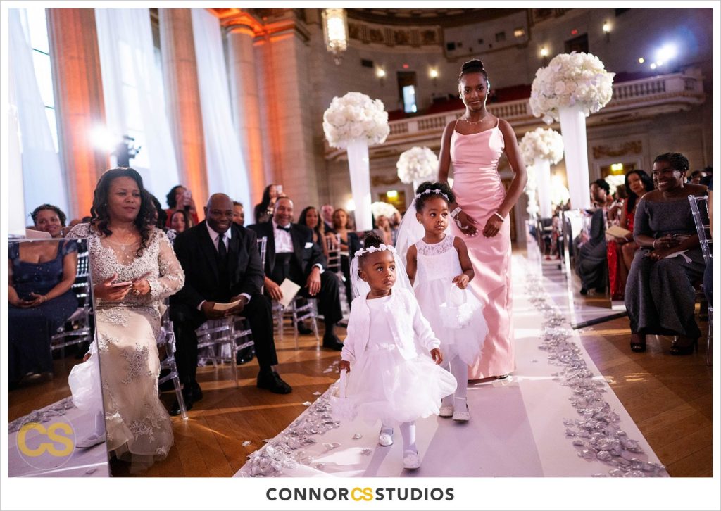 flower girls entering wedding at the Andrew W. Mellon Auditorium in Washington, DC by Connor Studios surrounded by tall white floral arrangements