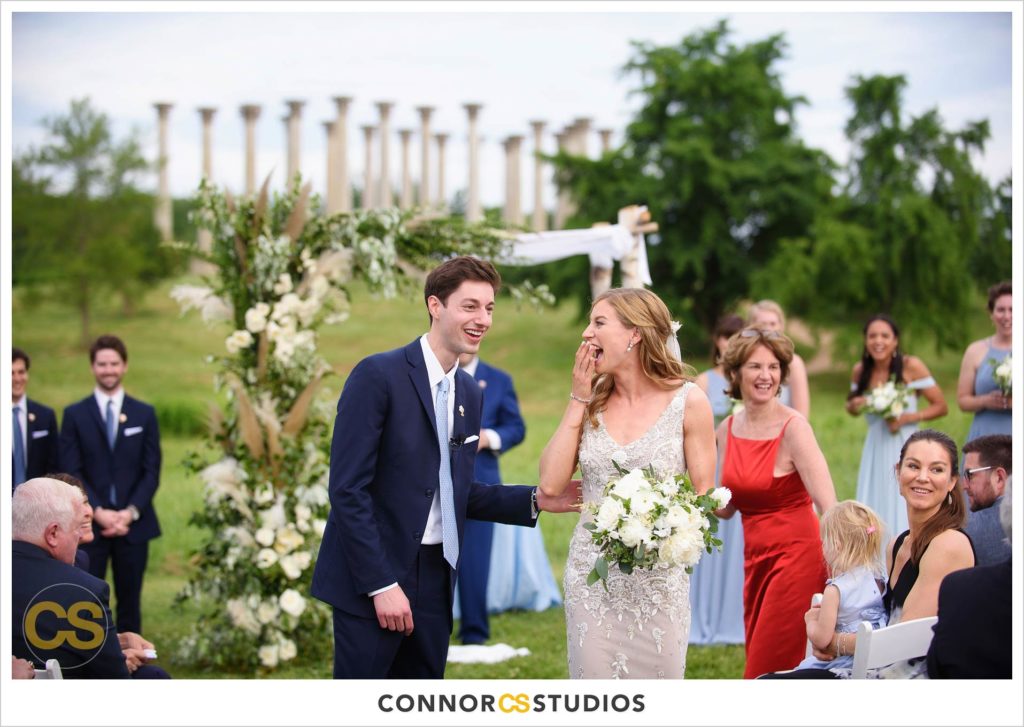 bride and groom walking down aisle during outdoor summer wedding ceremony with the National Capitol Columns in the background at the national arboretum in washington, dc by connor studios